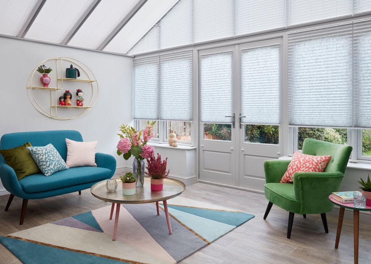 Conservatory with pale grey Pleated blinds on the side windows lowered three quarters and white Pleated roof blinds fully closed. Electric blue sofa, teal chair, round glass table on a blue and white geometric patterned rug.