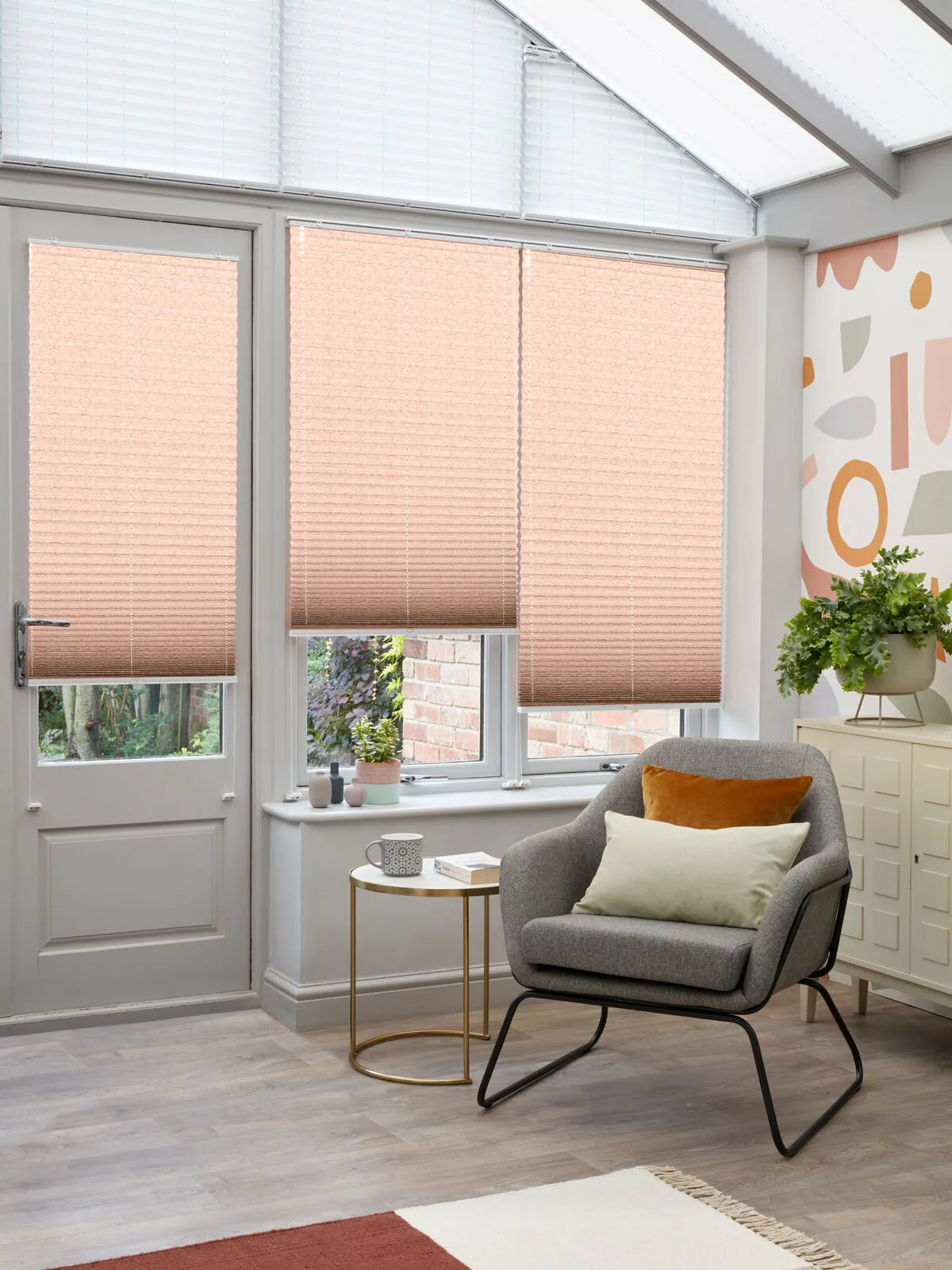 Conservatory with side windows dressed in rust coloured Pleated blinds, at varying heights, modern grey chair with cream cushion, russet rug on pale wood floor.