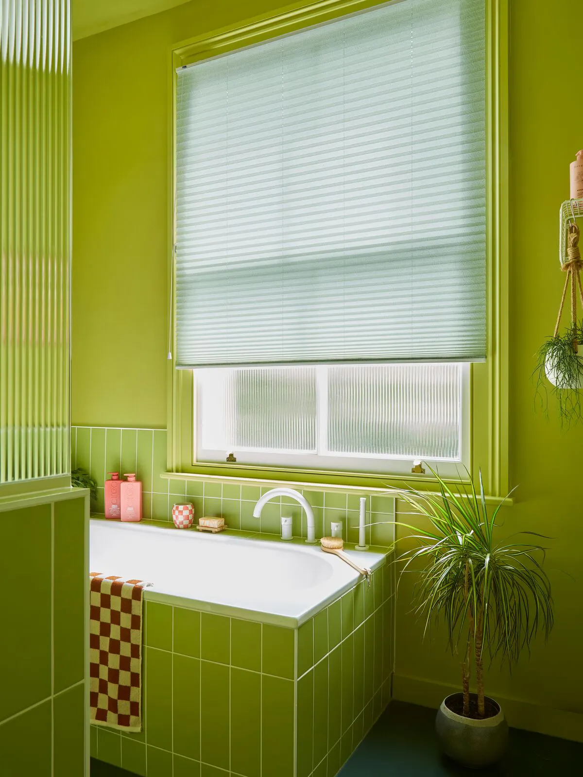 Retro-style bathroom in mid green with window dressed in grass green pleated blind, lowered three quarters, above a bath tiled to match the walls, potted plant alongside.