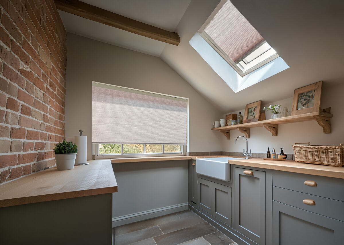 Soft grey kitchen with brick feature wall, pale wooden worktops and skylight and window fitted with light blush pleated blinds.