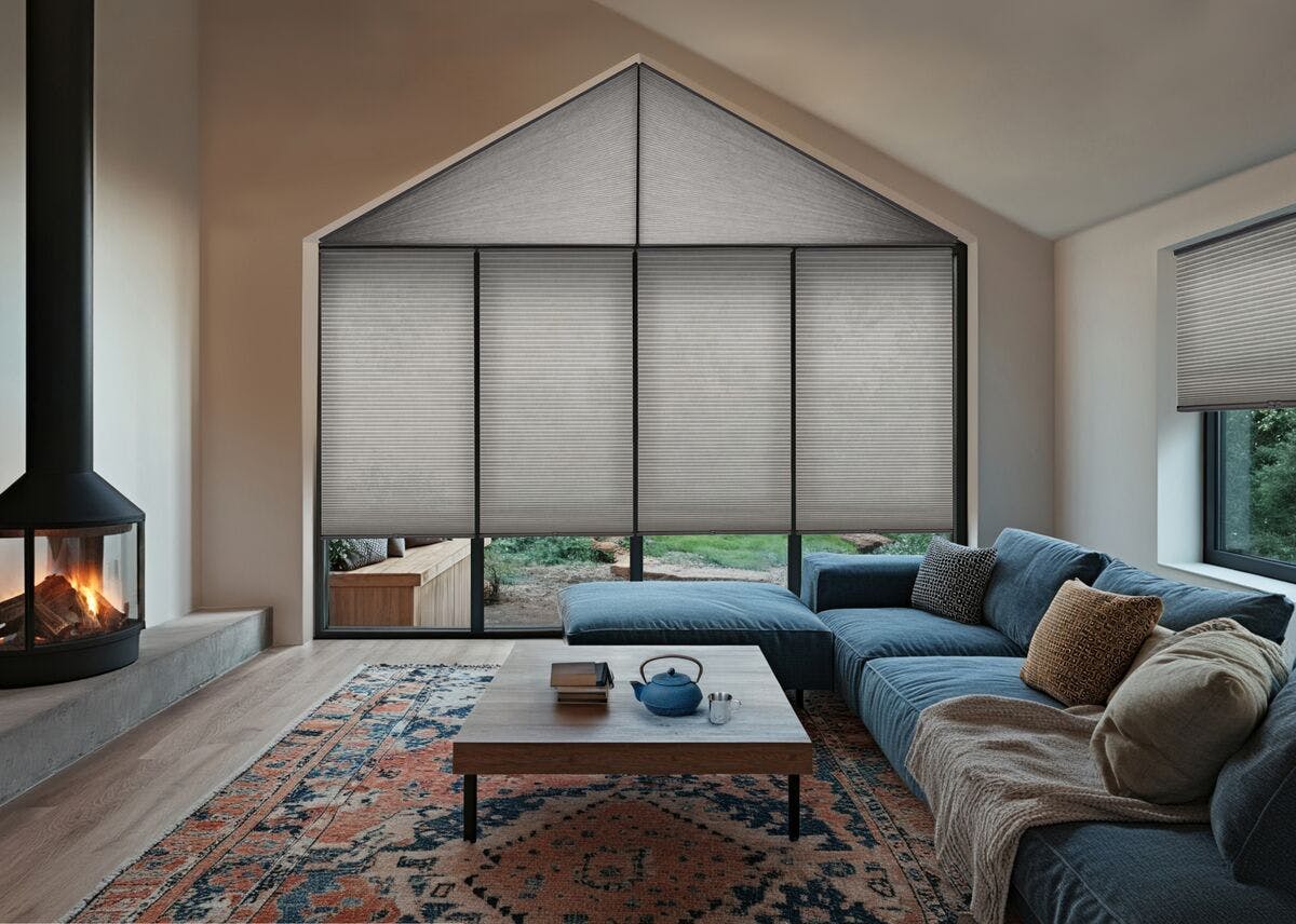 Neutral living room with large gable window and side window fitted with grey pleated blinds, warm tones and a blue sofa.