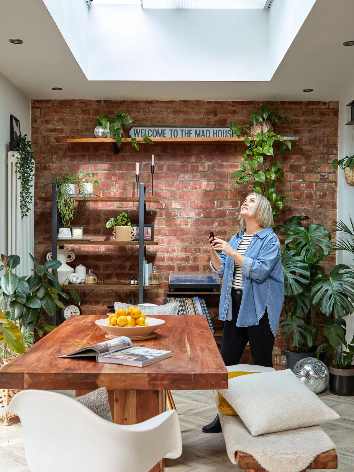 Bright dining space with a large skylight window and soft grey blind, exposed brick wall, green plants and warm wooden tones.
