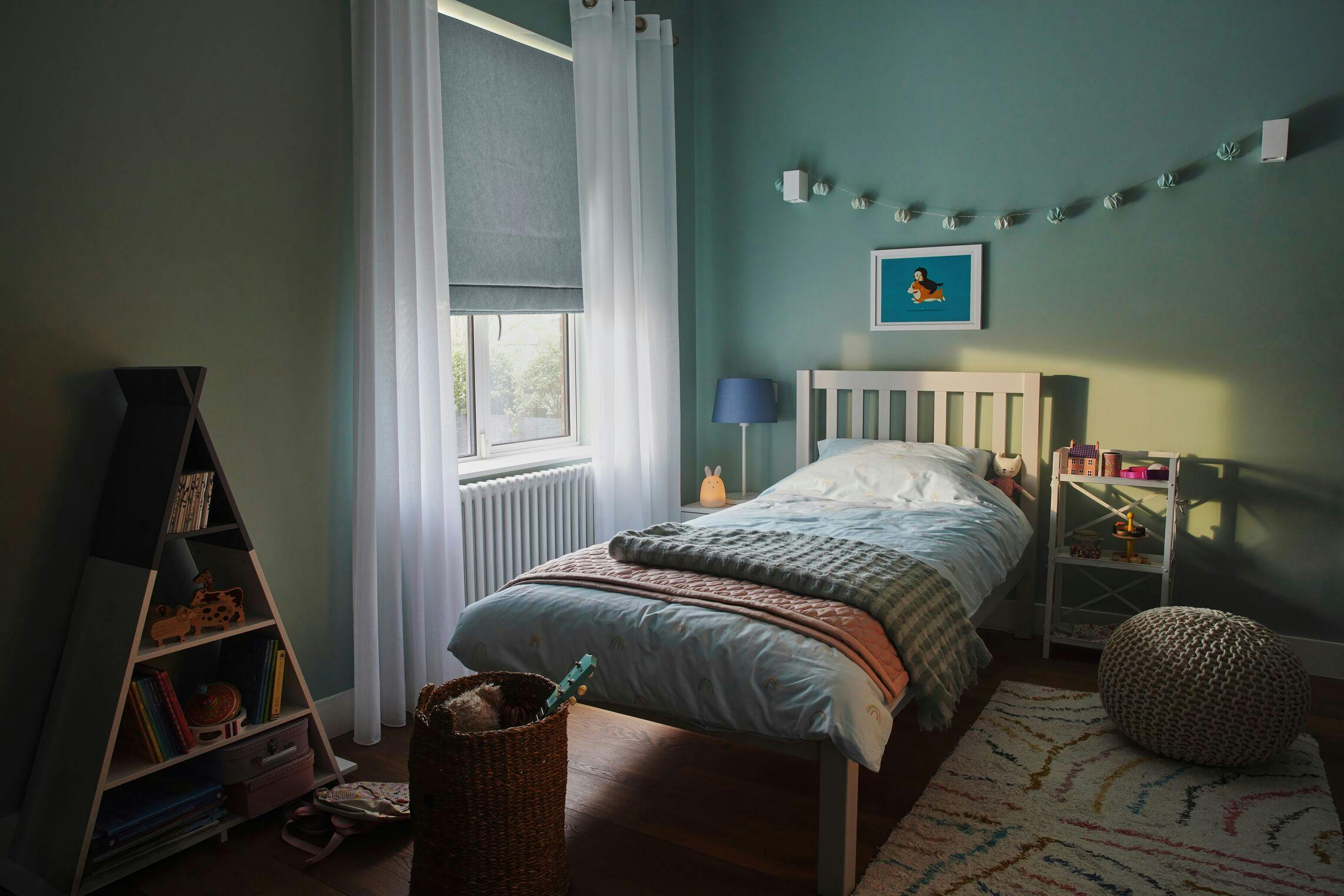 A small bed with pastel bedding is positioned near a window, surrounded by toys and books on shelves, in a sunlit, neatly organized children's room with teal walls and playful decor.