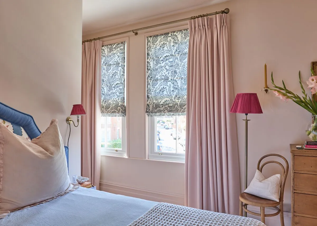 Bed with blue headboard and pillows beside a wooden drawer and chair. Vase of pink flowers and books atop drawer. Sunlit room features floral curtains and pink lamps.