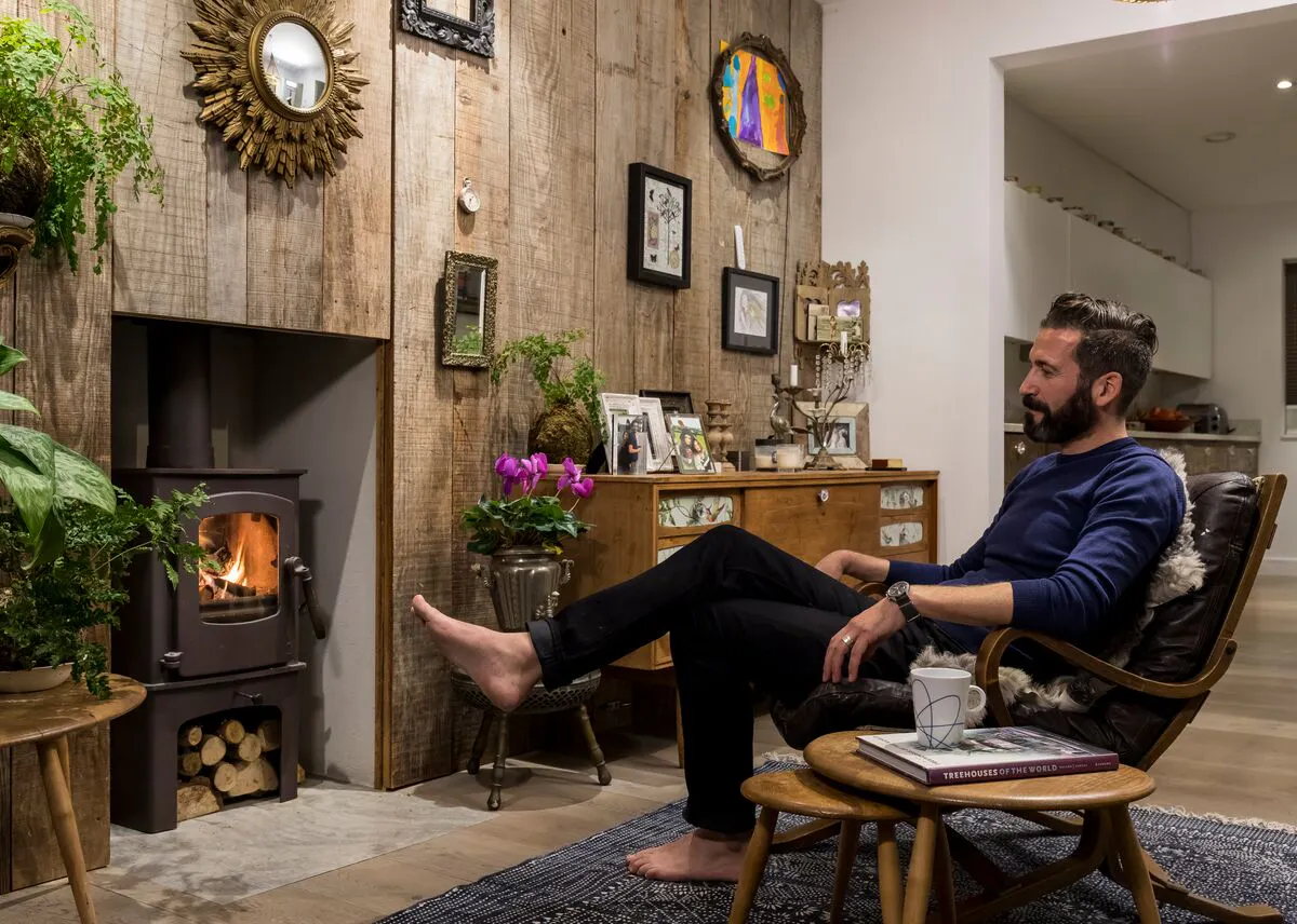 Oliver Heath sat in a cosy living room with a wood‑burning stove, rustic timber wall and eclectic décor.