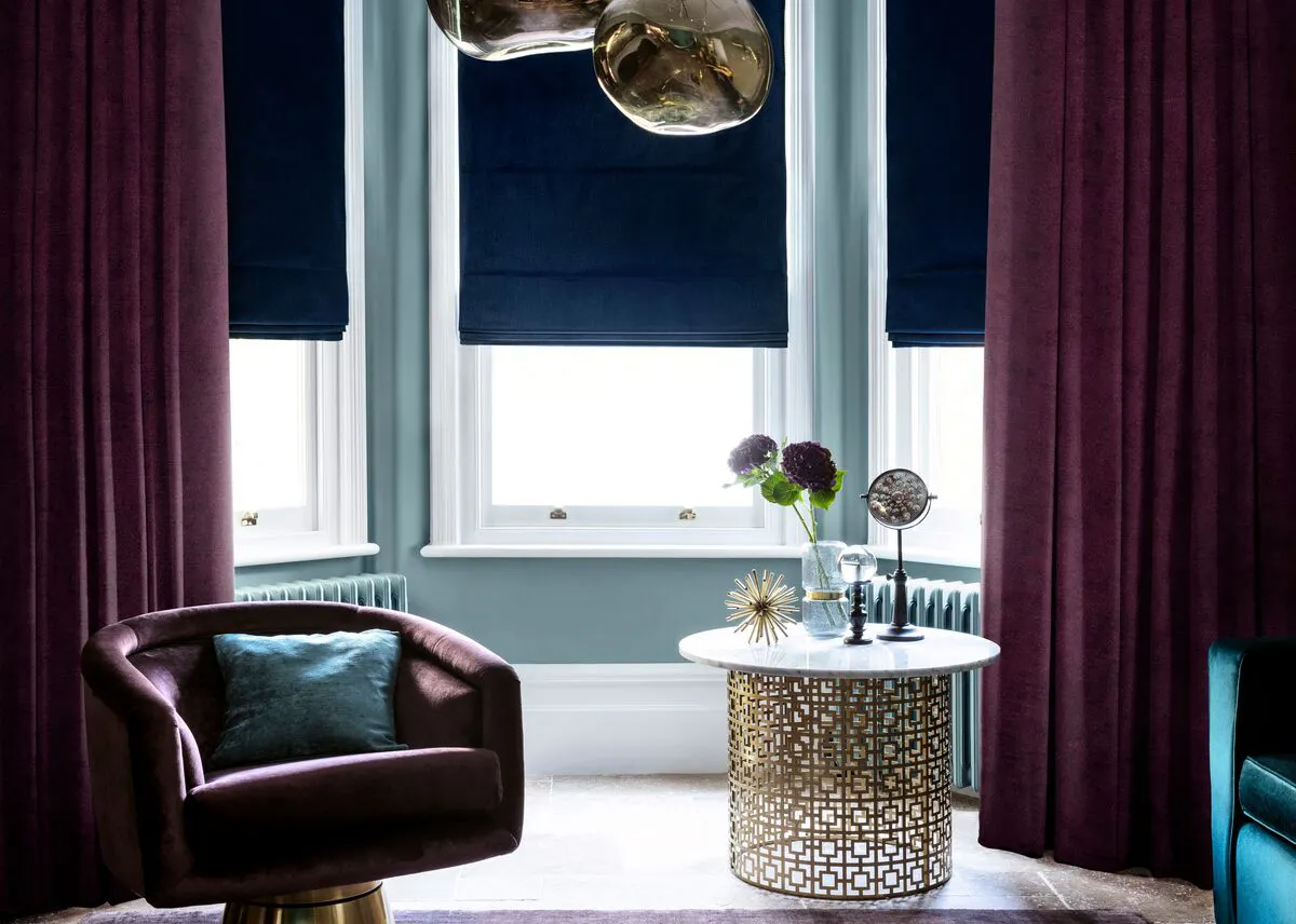 Bay window with deep blue Roman blinds framed by rich plum curtains, creating a moody lounge with soft light and layered textures.