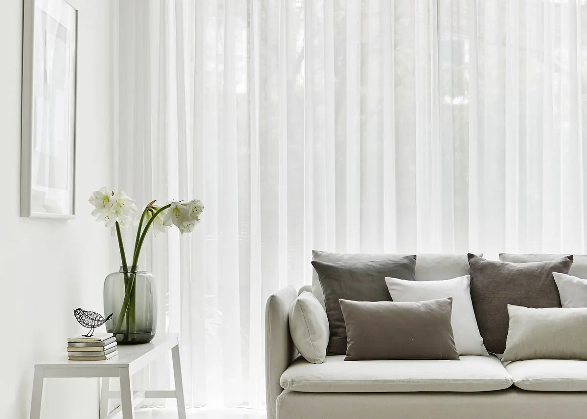 Sheer voile curtains filter daylight through a wide living room window, creating a bright, airy feel beside a neutral sofa and soft furnishings.

