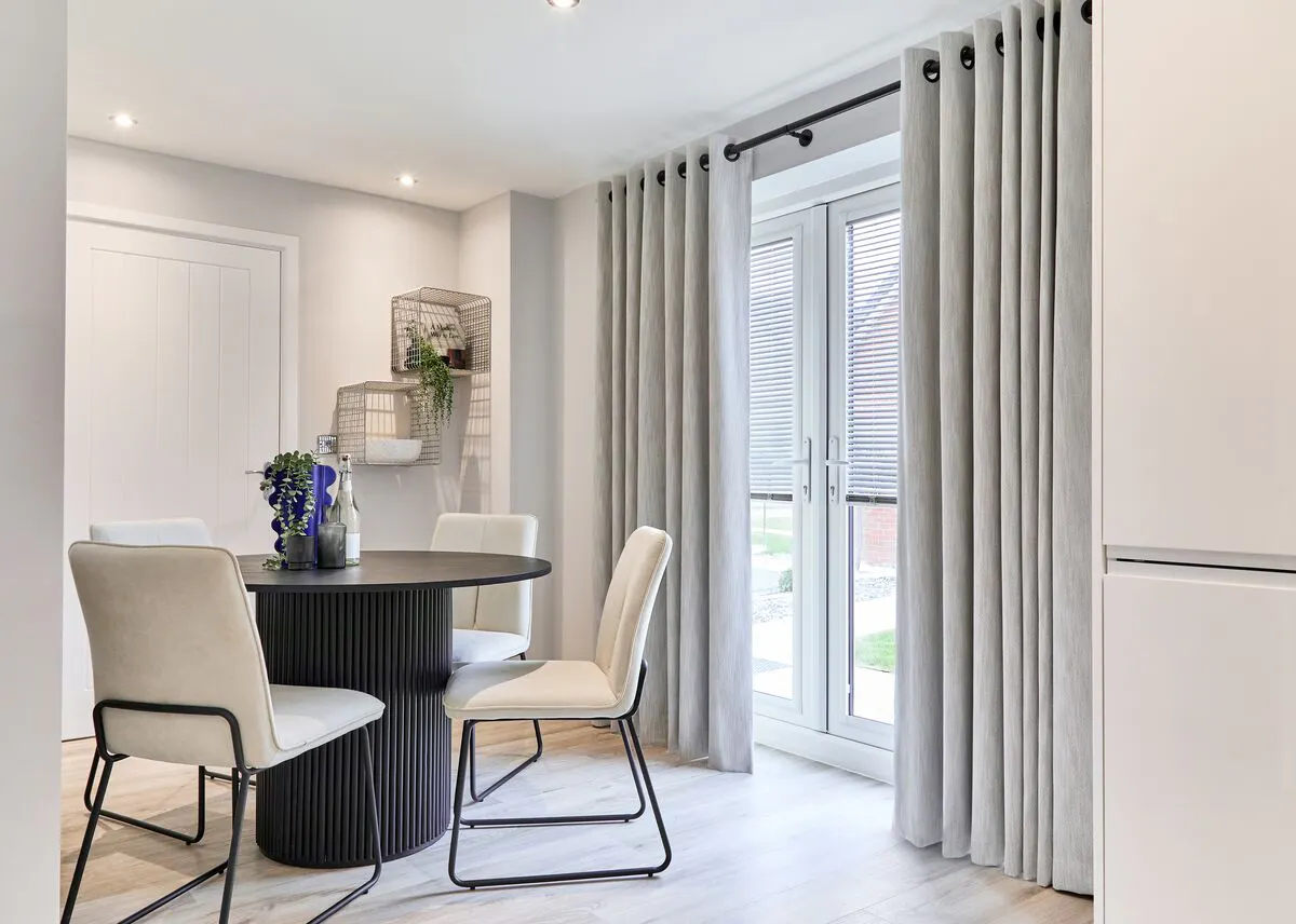 Grey eyelet curtains frame French doors in a modern dining area, letting natural light in beside a round table and upholstered chairs.