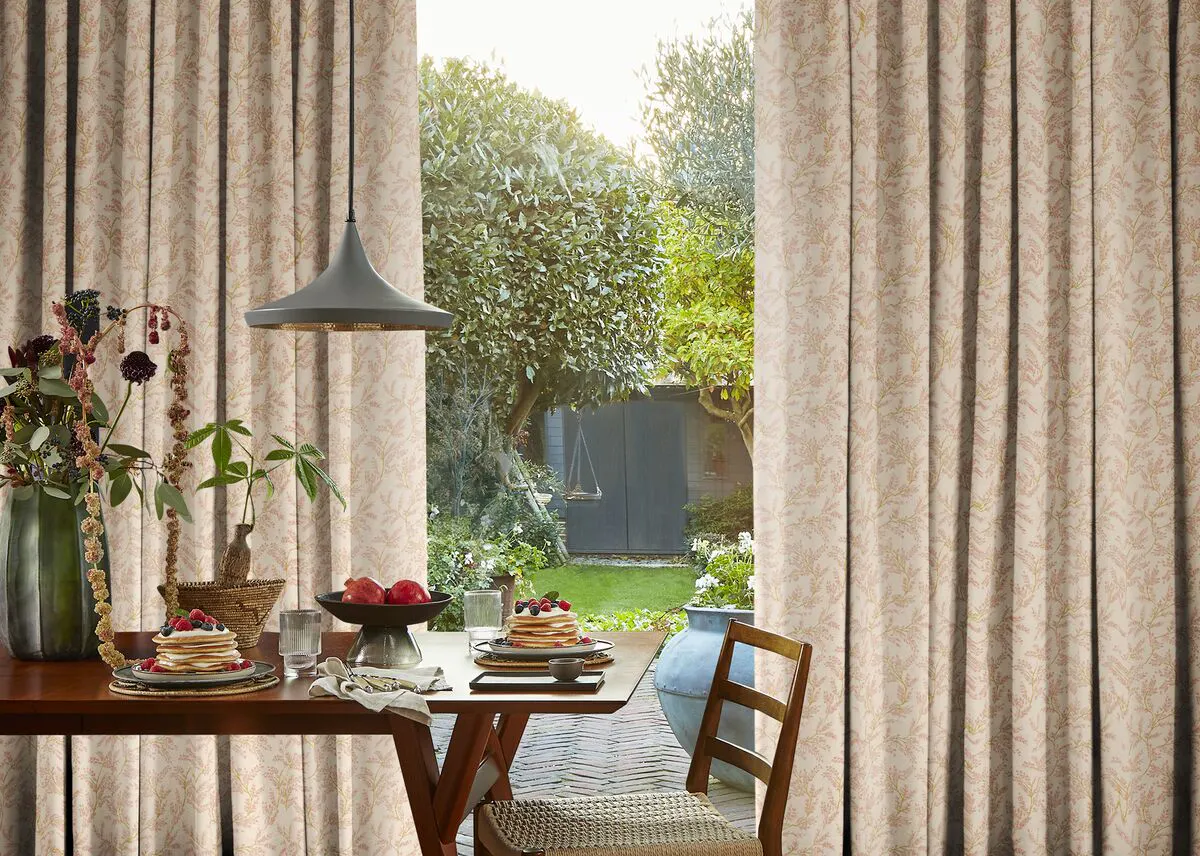 Patterned curtains hang either side of a wide patio window, opening to garden views and soft daylight in a cosy dining space.