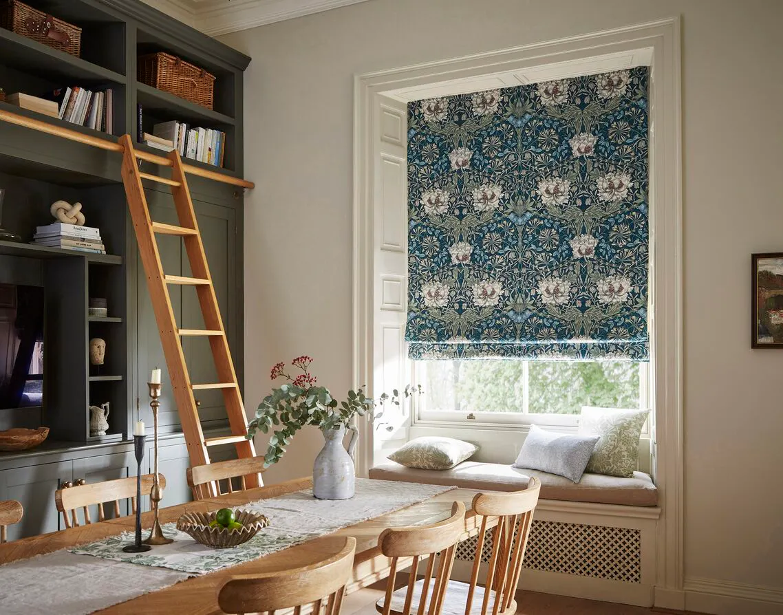 Dining room window dressed with a patterned Roman blind, framed by white trim, with a window seat beneath and soft daylight filtering through.