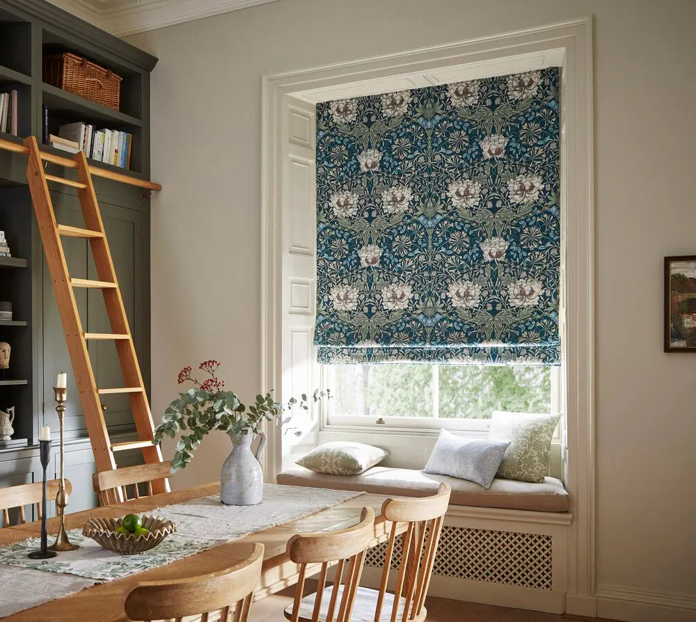 Dining room window dressed with a patterned Roman blind, framed by white trim, with a window seat beneath and soft daylight filtering through.