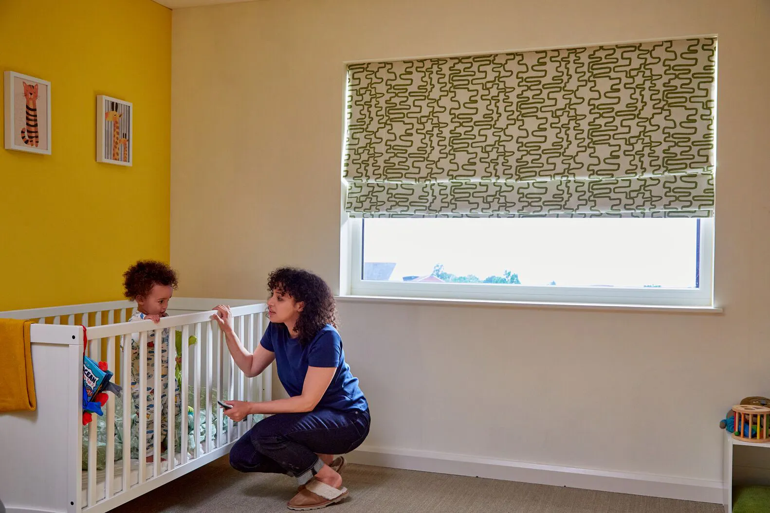 An adult leans beside a child’s cot in a bright room with a patterned Roman blind over a large window.