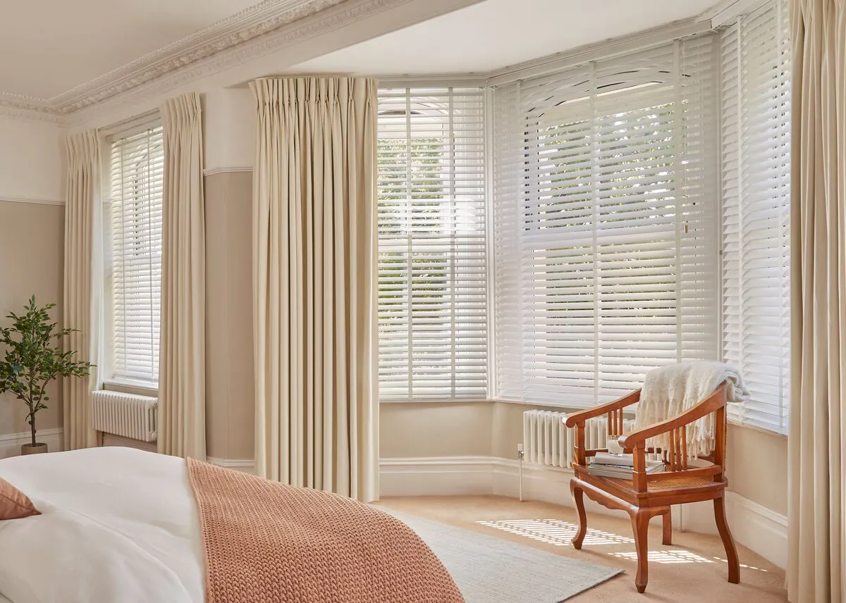 A bright bedroom featuring tall cream curtains framing wide windows with white blinds, filling the space with natural light.