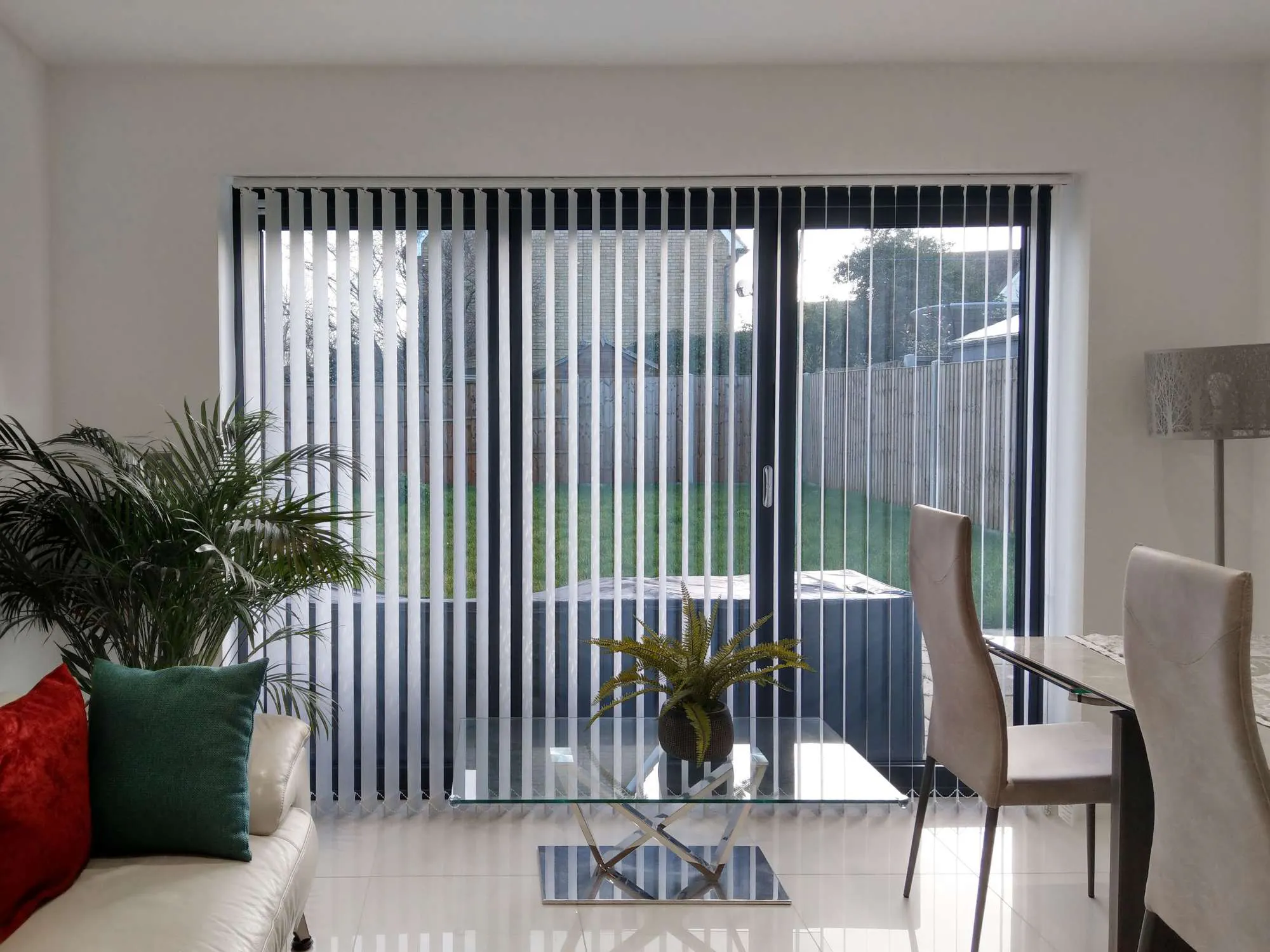 Vertical blinds cover glass doors, partially opened to a backyard. A cream sofa with red and green pillows sits nearby, accompanied by a glass coffee table with a plant, in a modern room.