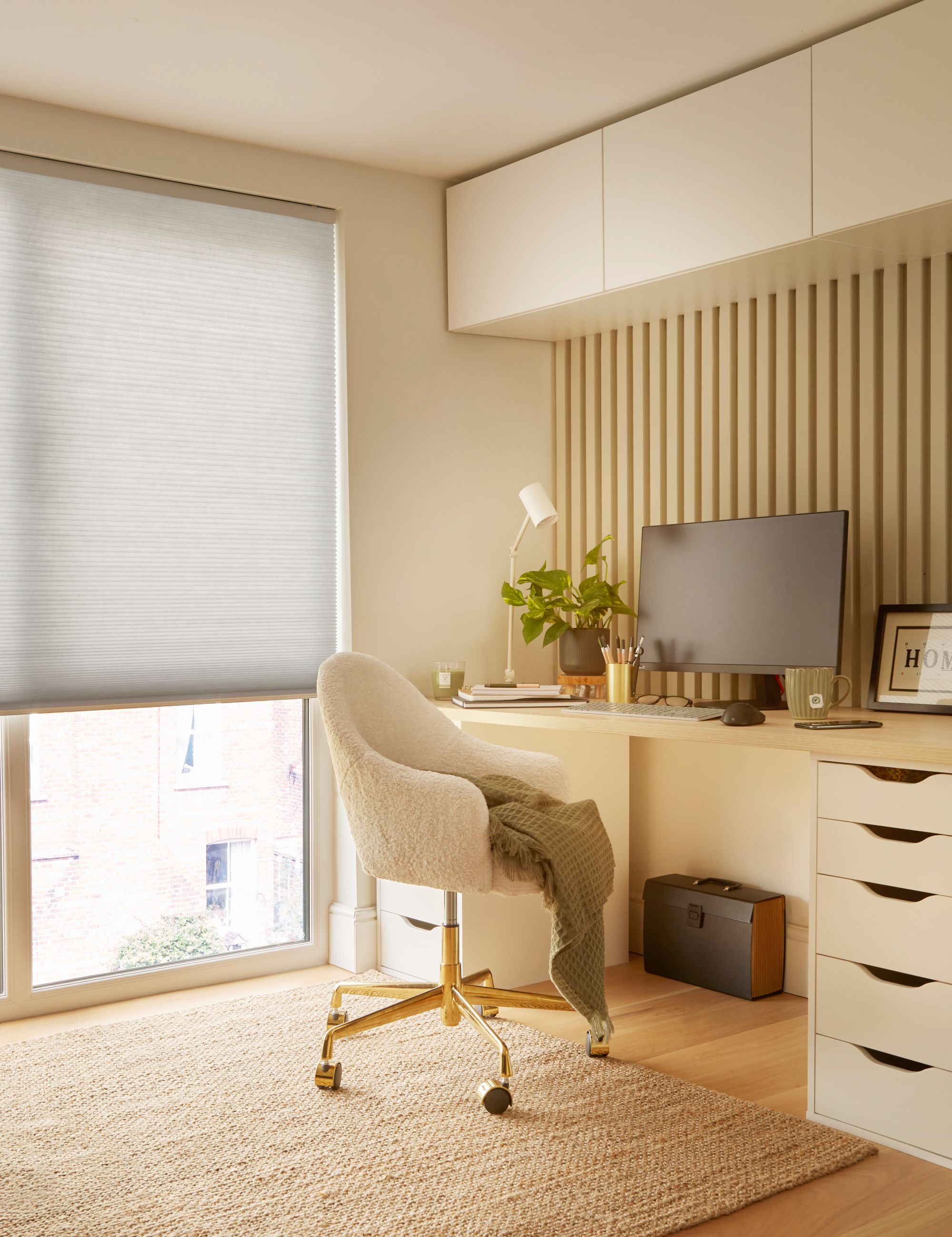 Ivory home office with floor to ceiling window dressed in pale grey duette blind, lowered halfway, ivory desk with drawers, high units above, matching chair, jute rug on wooden floor, monitor, small plant and desk lamp.