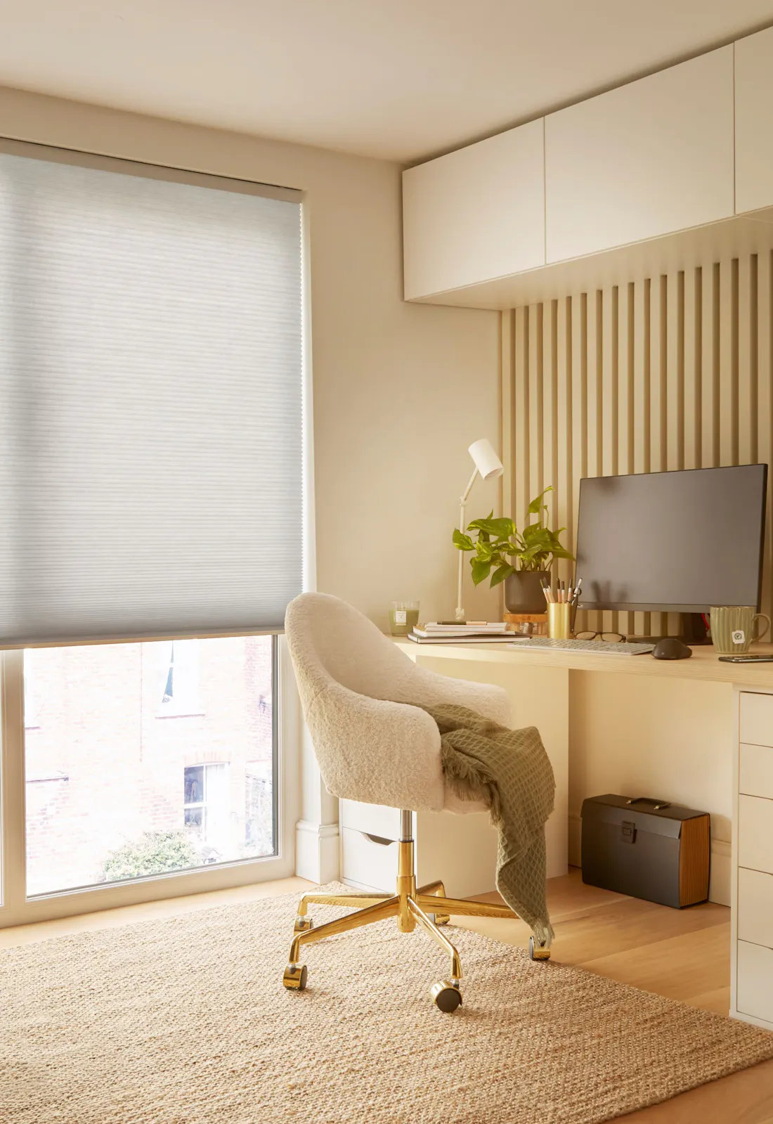 Ivory home office with floor to ceiling window dressed in pale grey duette blind, lowered halfway, ivory desk with drawers, high units above, matching chair, jute rug on wooden floor, monitor, small plant and desk lamp.