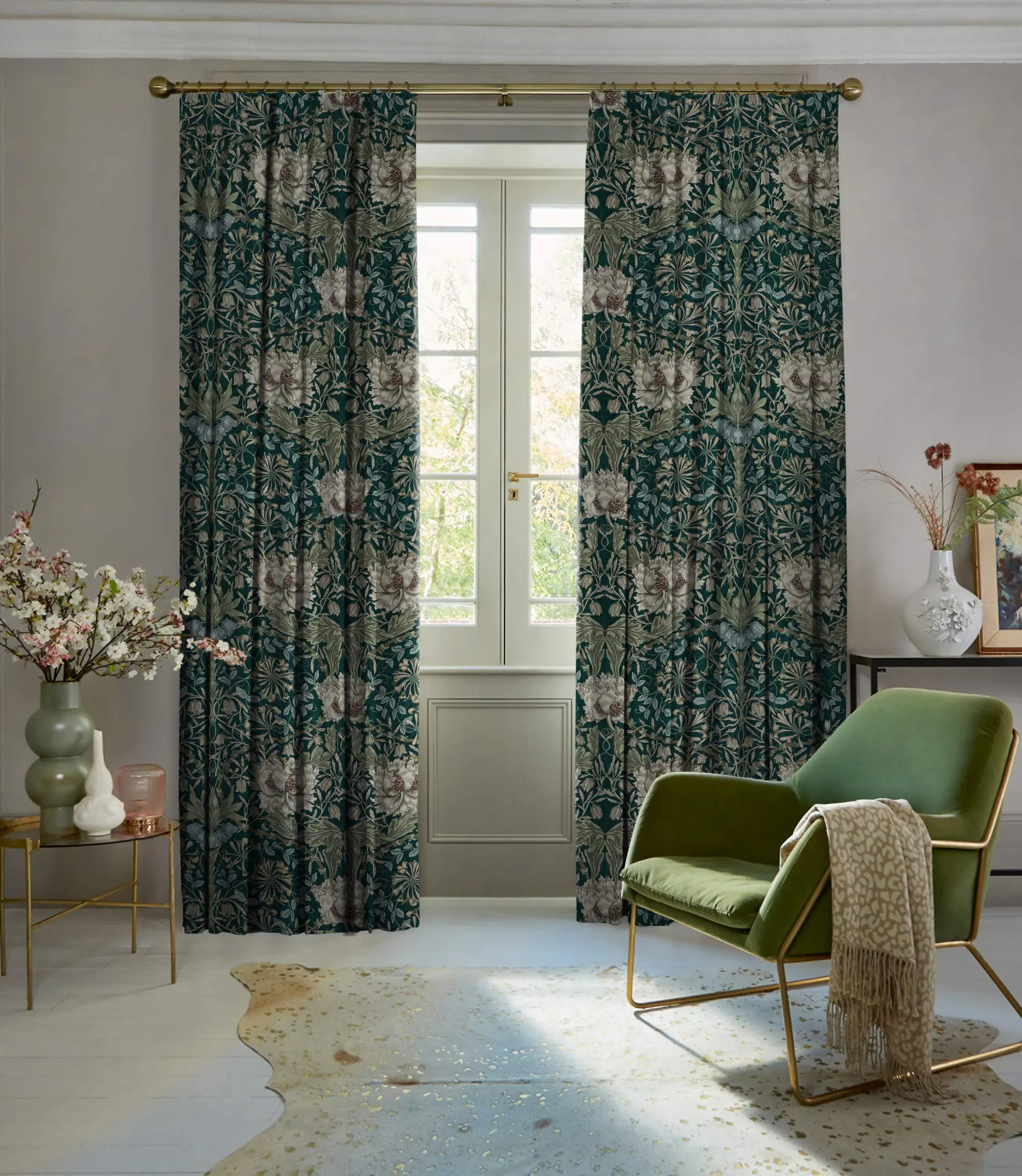 Living room in pale mink with curtains in dark green with cream floral pattern, partly open, polished concrete floor, green velvet occasional chair, with pale mink throw over arm, round table with green vase with foliage.