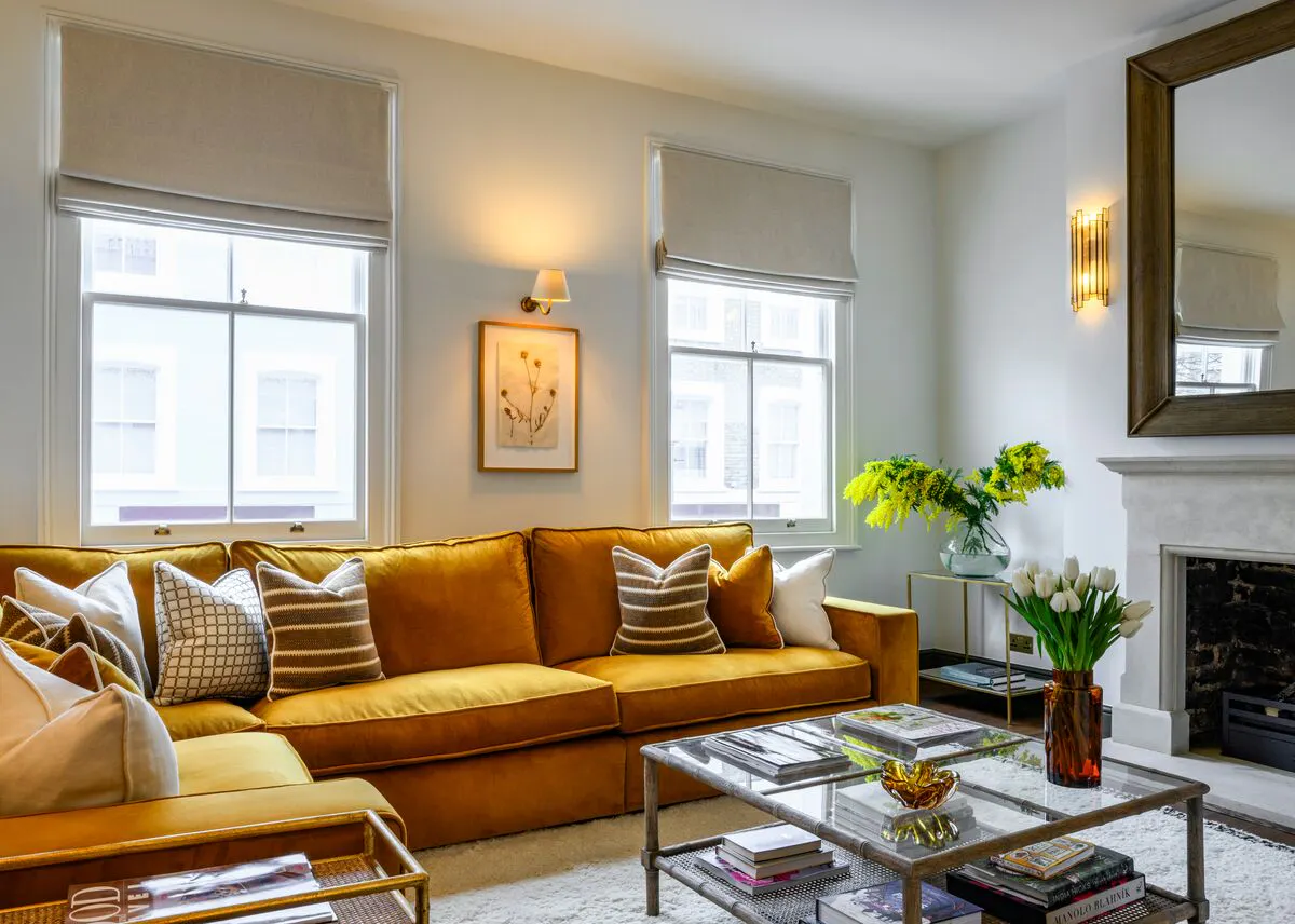 A bright living room with two windows dressed in pale Roman blinds, allowing soft daylight to filter through above a mustard sofa.