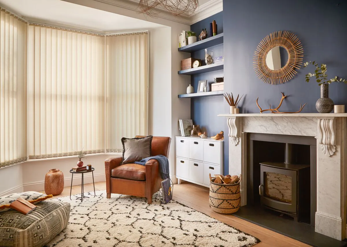 A bay window with cream vertical blinds lighting a cosy room with a leather armchair, rug and a marble fireplace.