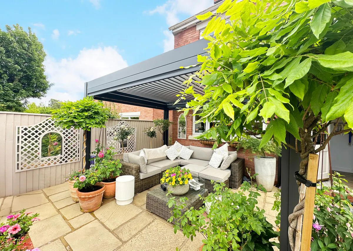 Sleek pergola shading a garden seating area, with rattan sofas, pale cushions and lush planting arranged across a sunlit stone patio.