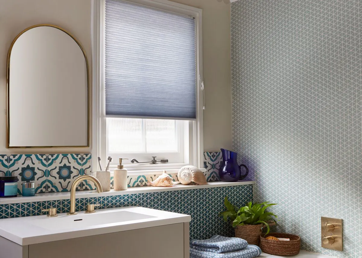 Bathroom window dressed with a soft blue blind above a vanity, framed by patterned tiles, with light filtering in over the sink and bath area.