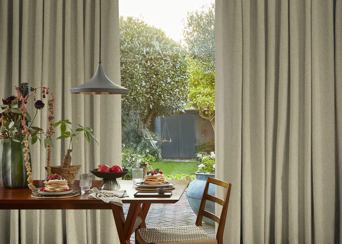 Dining room with full size window and doors dressed in pale taupe pinch pleat curtains, partially drawn, wooden table, chairs and bench flowers in vase.