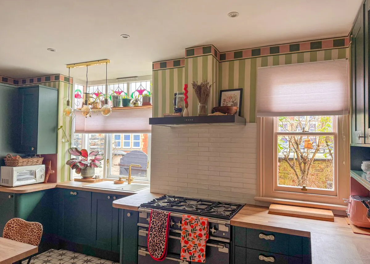 A pair of windows with Honeycomb blinds bringing light into a patterned kitchen with green cabinets and wooden worktops.