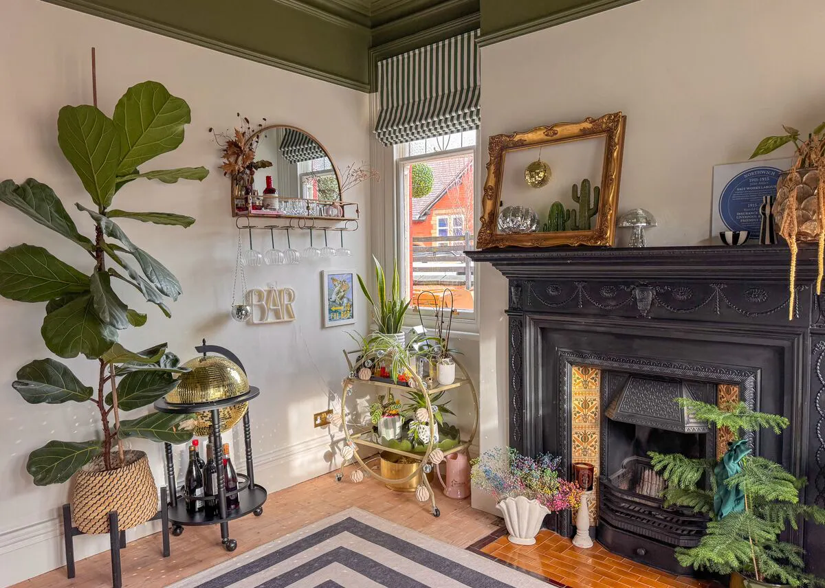 A small window with a striped Roman blind sits beside a dark fireplace, surrounded by plants and eclectic décor.