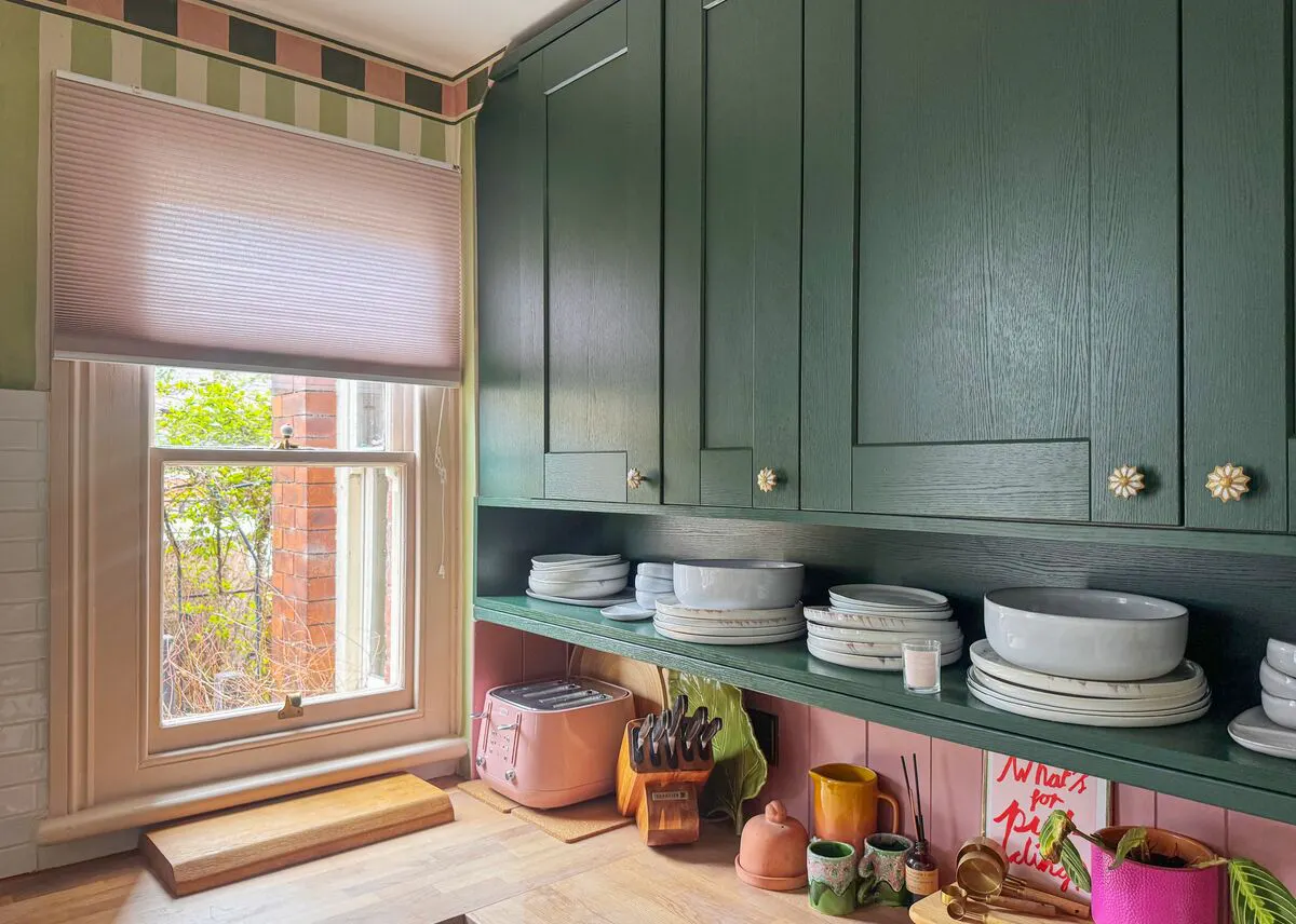 A window with a Pleated blind brings light into a kitchen corner lined with green cabinets, open shelving and stacked pottery.