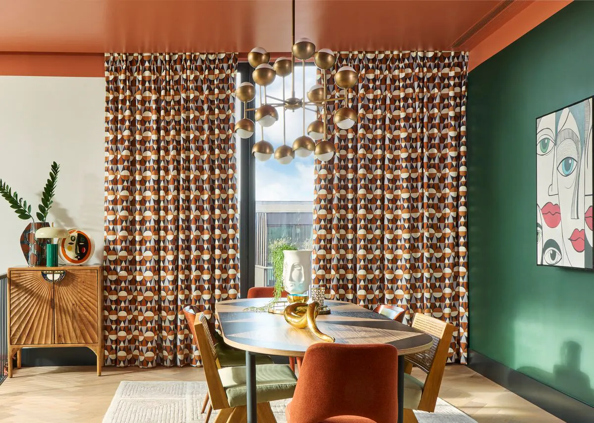 A vibrant dining room with tall windows dressed in bold patterned curtains, framing a view of the outdoors behind the round table. 