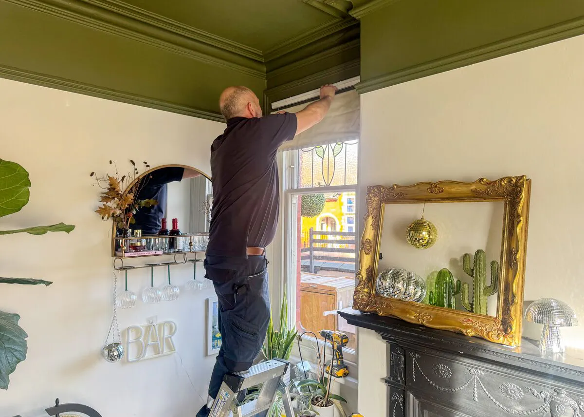 Blind being fitted above a tall window in a characterful living space, with ornate cornicing, plants and a decorative mirror framing the area. 
