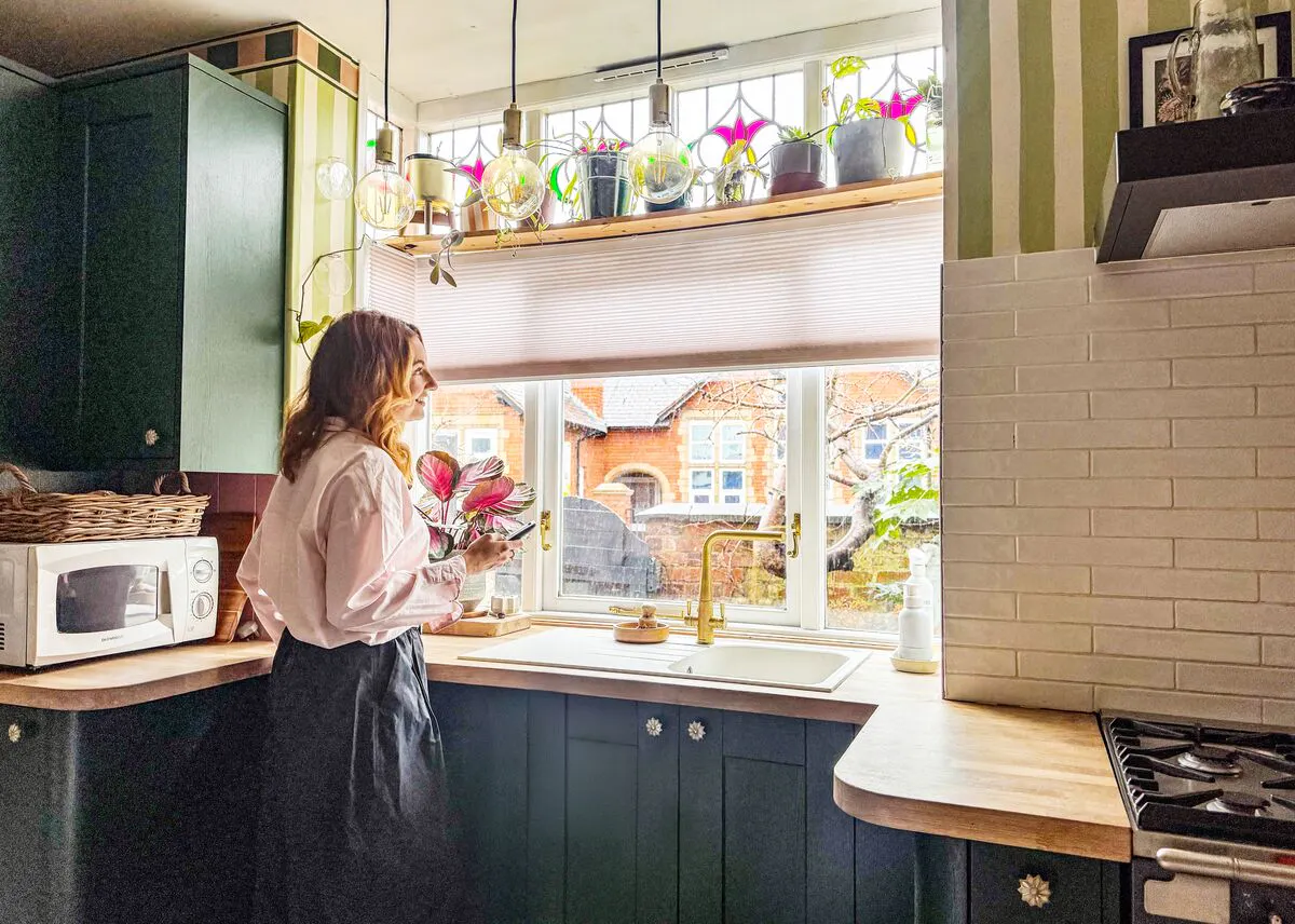 Person standing at a kitchen sink beside a wide window with a Pleated blind half lowered, plants on the sill and daylight filling the green kitchen.