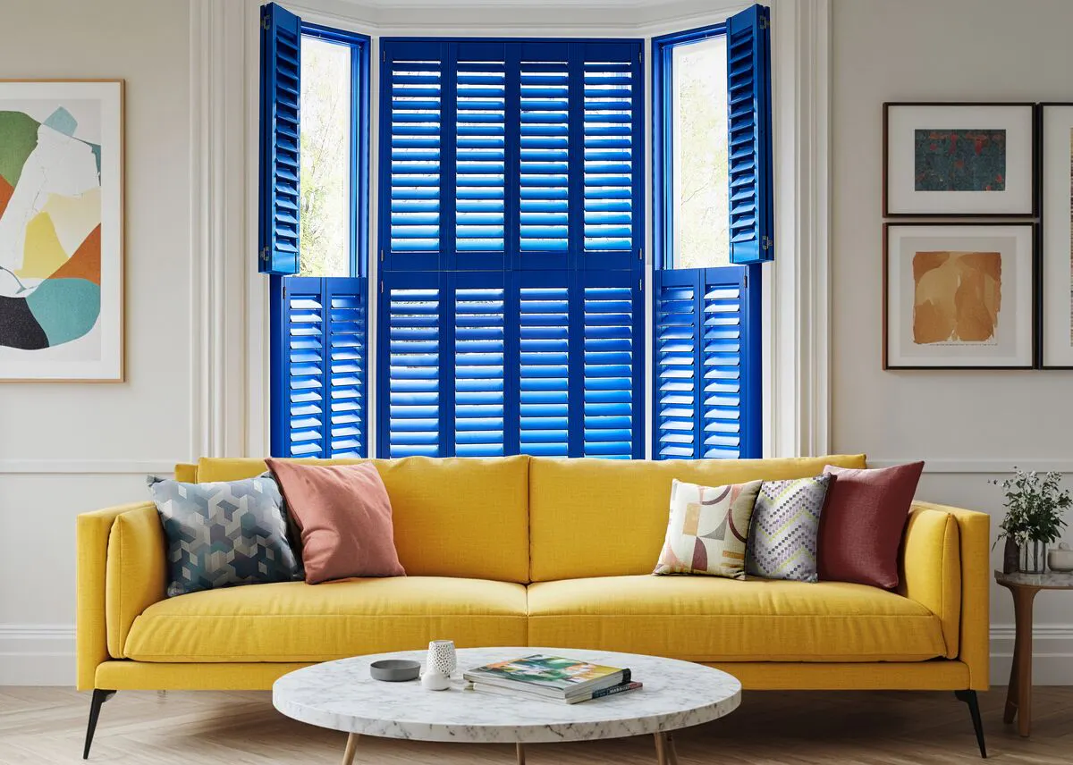 Bright living room with a bay window dressed in bold blue shutters, paired with a yellow sofa and colourful cushions.
