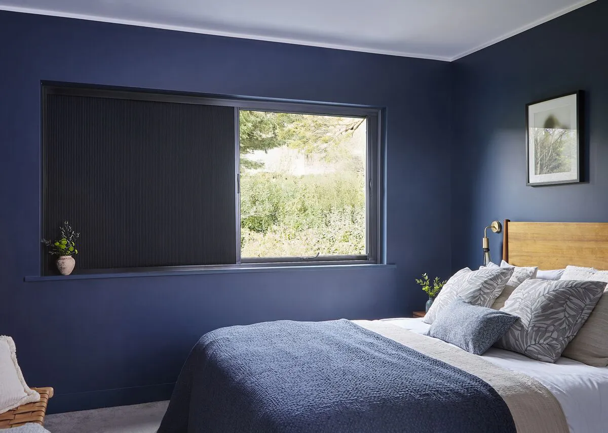 A dark blue bedroom with a black Blind Screen at the window, allowing filtered light and a view of greenery outside.