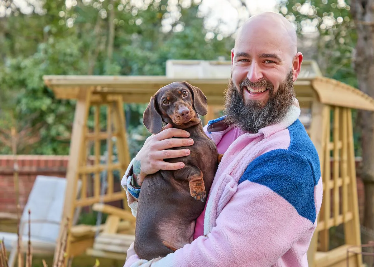 A person outdoors holding a small brown dachshund, with a wooden garden swing and foliage in the background. 