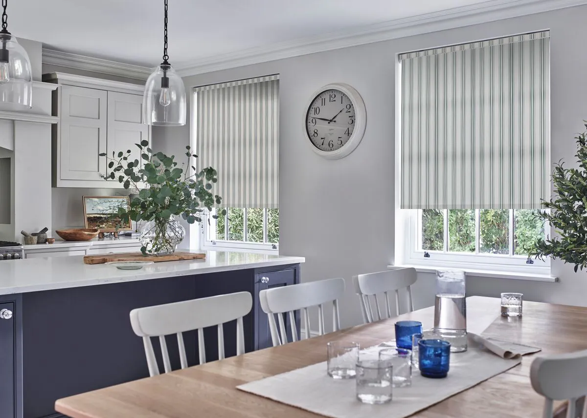 A bright kitchen-diner with two windows fitted with soft striped blinds, bringing light into the space above pale walls and a wooden table.
