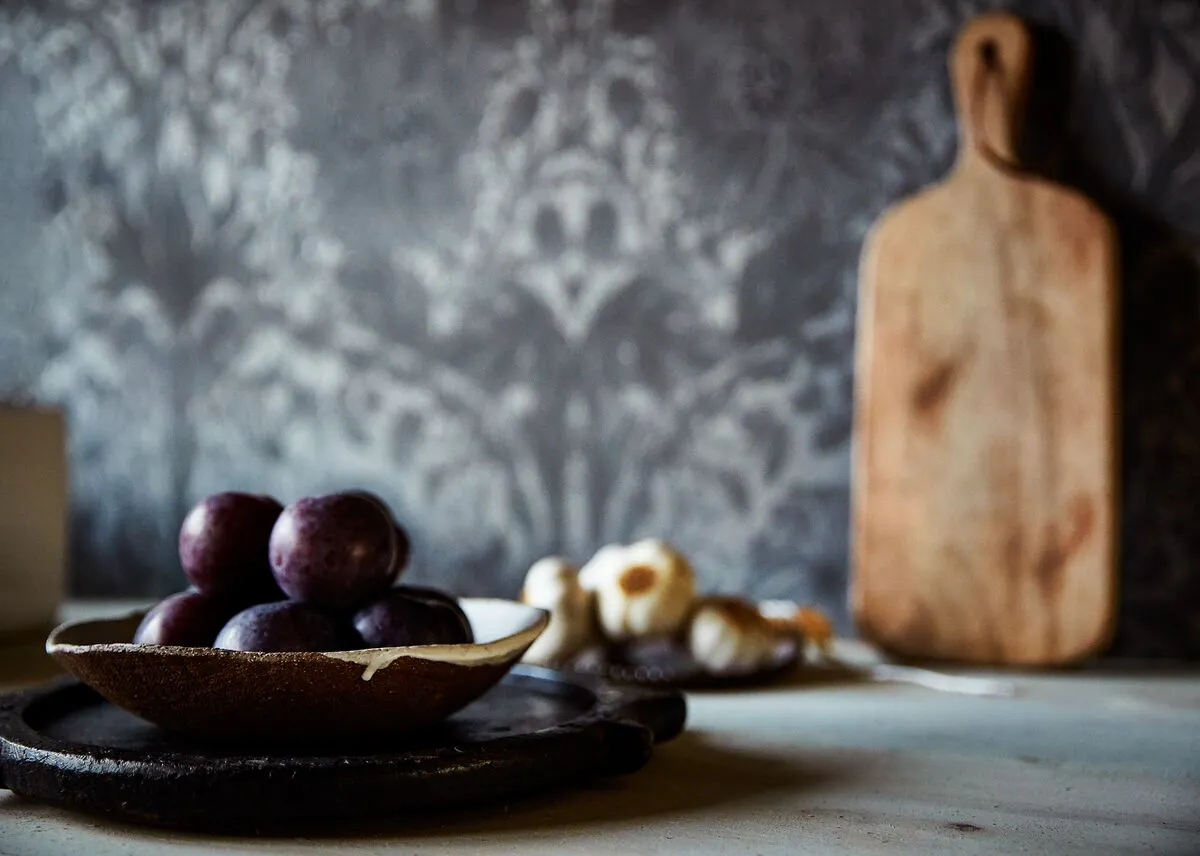 A rustic kitchen scene with a bowl of dark plums in the foreground, garlic and a wooden chopping board blurred in the background.