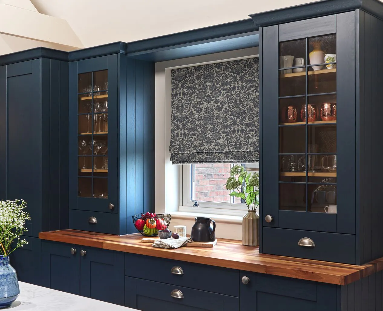 A small kitchen window dressed with a patterned Roman blind, set between dark blue cabinets with wooden worktops.