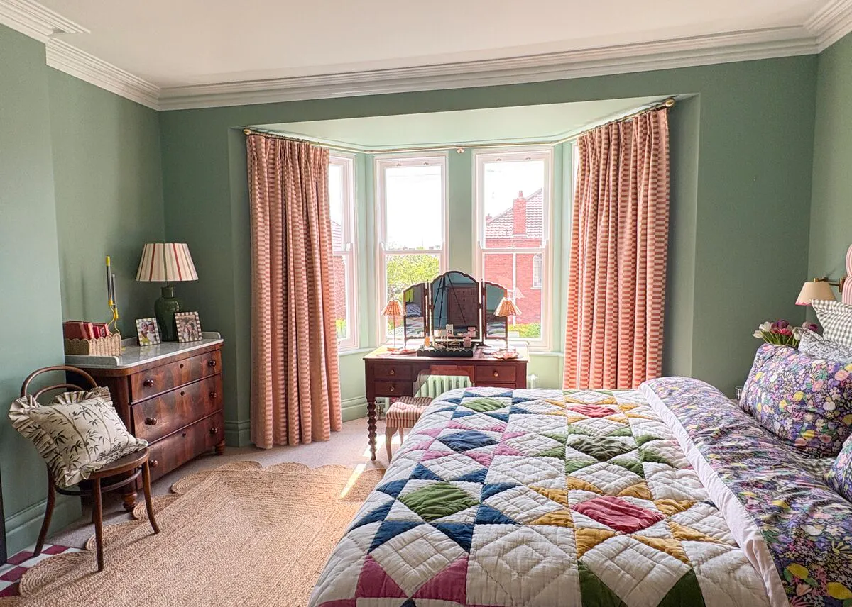 Pale green bedroom with deep recess bay window with curtains in pale salmon with small copper rectangle, open, bed with pinstriped headboard and handmade-style quilt  with accent colours in copper, blue and green reflected in mirror of dressing table in bay.