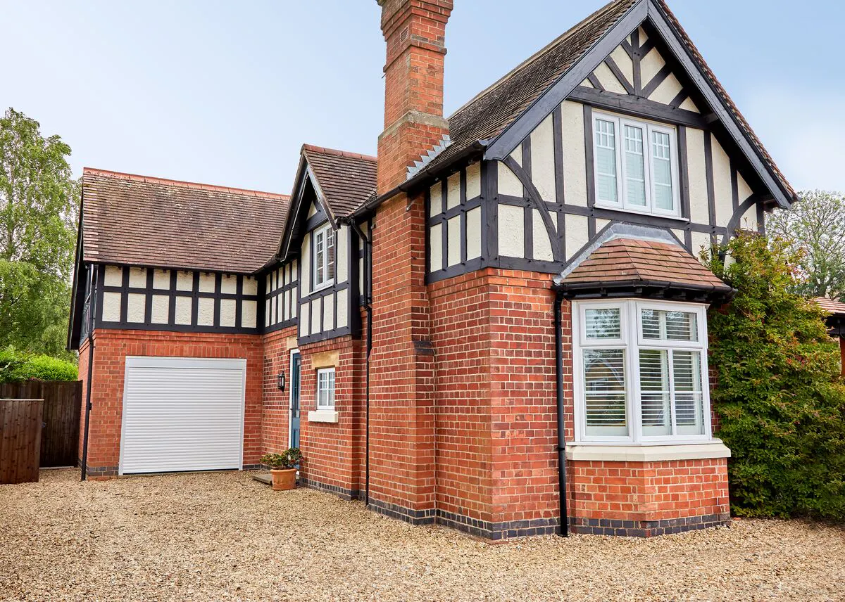 Exterior Tudor-style house in red brick with integral garage with white roller door, shingle all around house and drive.