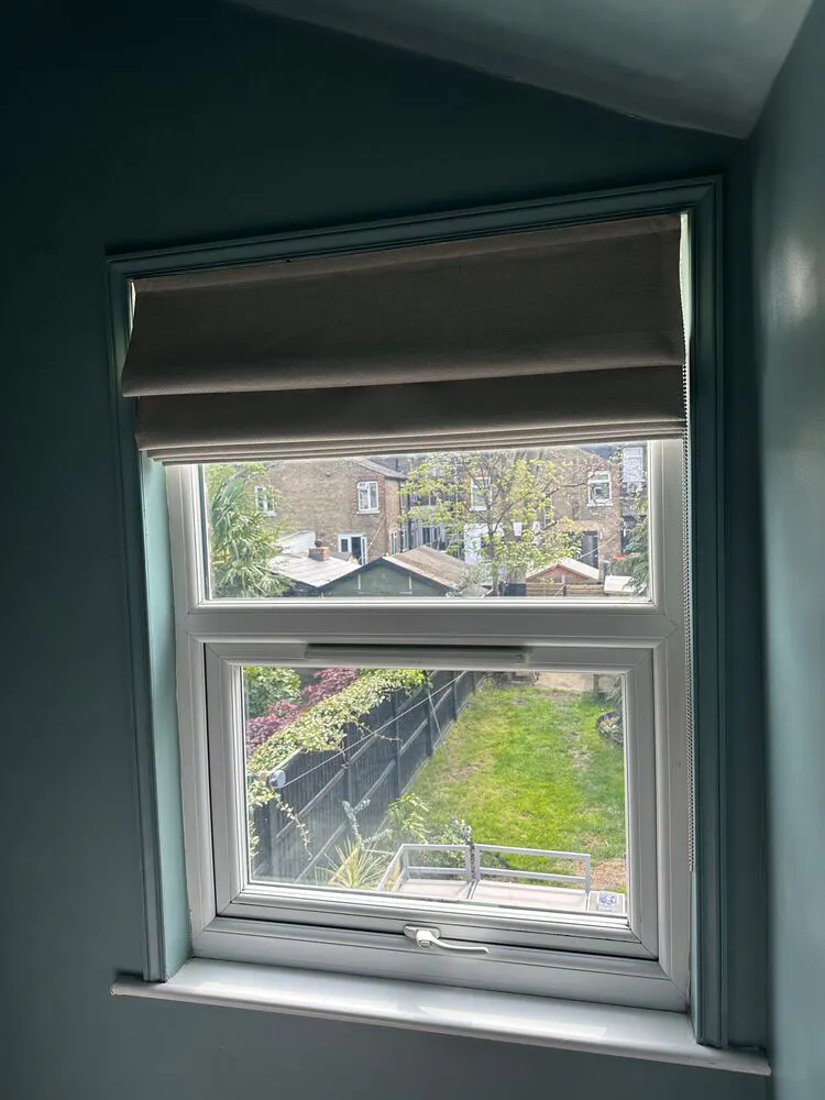 Small window in blue room dressed with mid grey Roman blind, raised, with view of garden below.