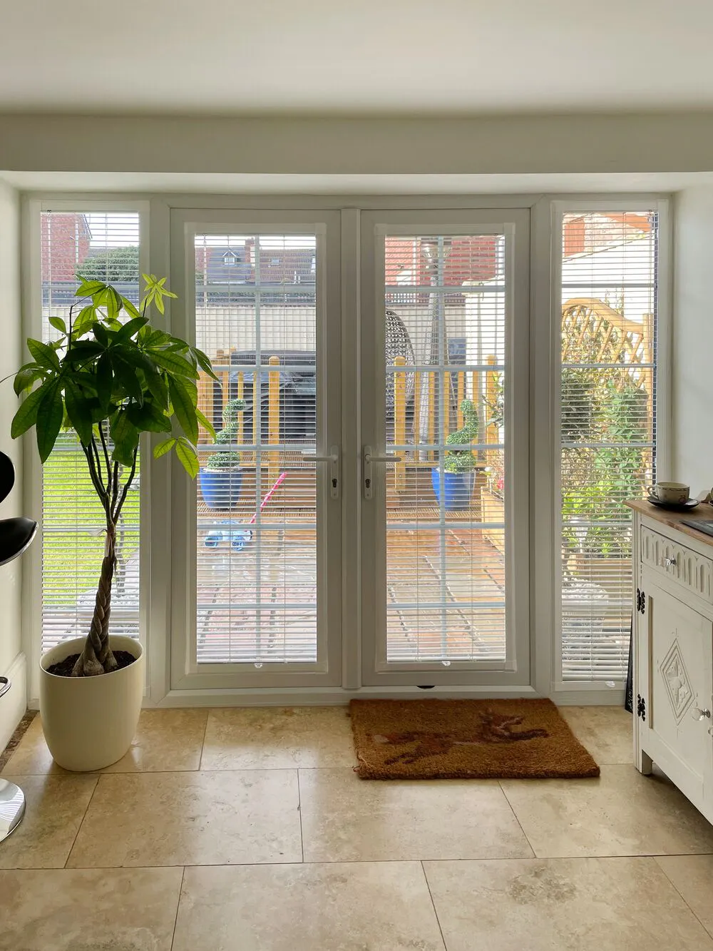 Cream kitchen with French doors and windows dressed in perfect fit cream venetian blinds, small tree in cream pot, coir doormat.