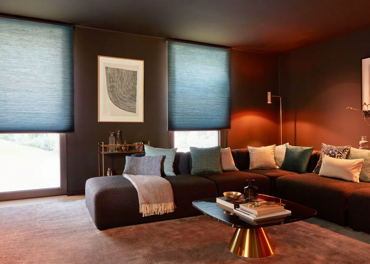 Living room with two blue blinds lowered over large windows, soft lighting highlighting a dark corner sofa and gold‑accented coffee table.