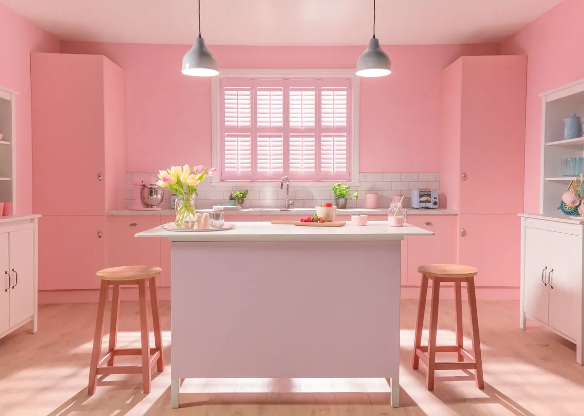 Pink kitchen with a central island and stools, featuring a wide window with shutters letting soft daylight brighten the space.
