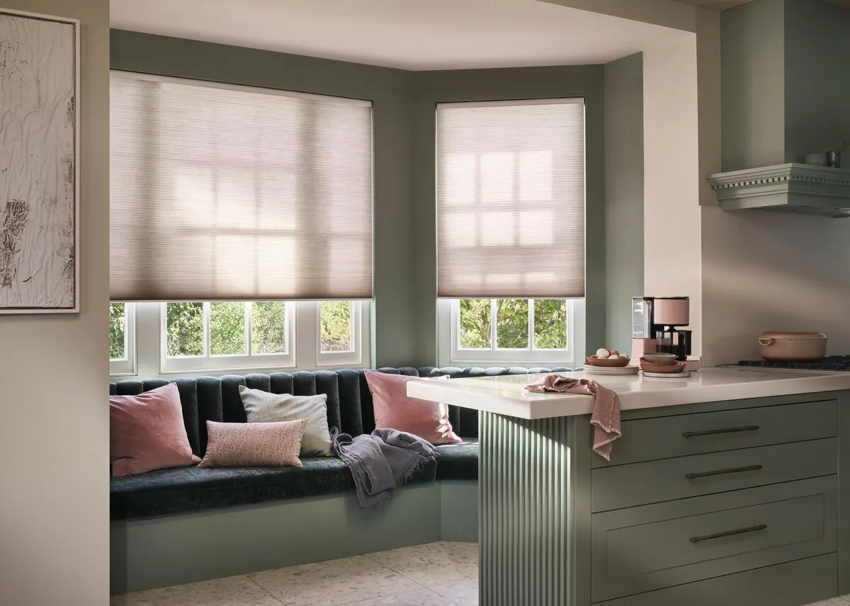 Built-in kitchen seating beneath a bay window, with soft Pleated blinds diffusing daylight across sage cabinetry and cushions.