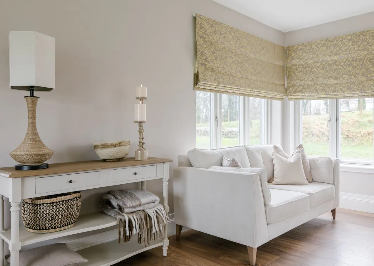 Cream living room with corner windows dressed in ivory patterned Roman blinds partially lowered, white sofa in corner, white console table with earthenware lamp with white square shade, candlestick and throw, wooden floor.