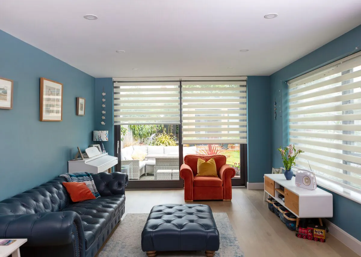 Living room with mid blue walls and white ceiling, window on right and patio doors to rear all dressed in white day and night blinds at varying heights, navy blue chesterfield sofa and footstool, wood effect flooring, rust-coloured armchair.