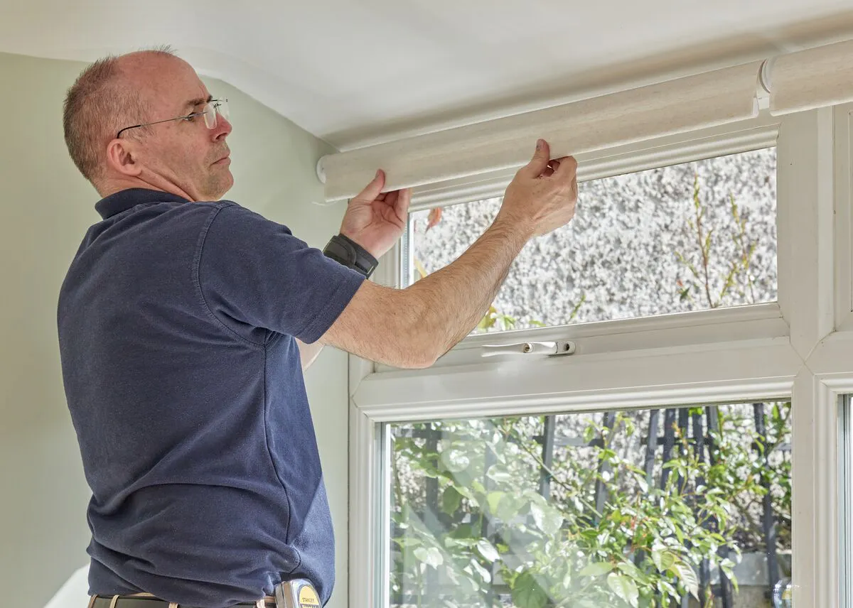 A man puts a Roller blind into a fitting above the top window in a conservatory.