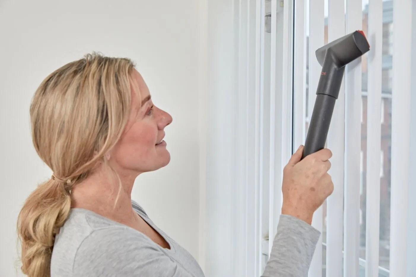 A woman vacuum cleans hanging blinds.