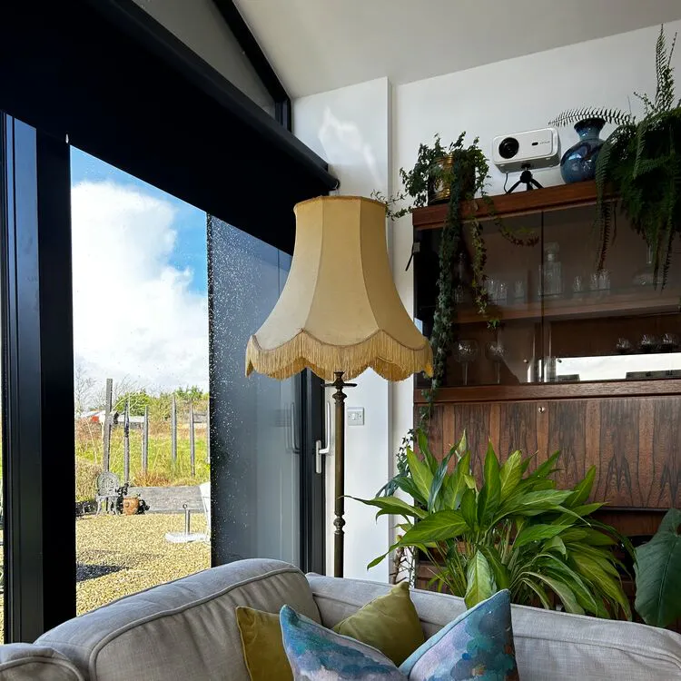 Close in on living room corner with patio door, brown sofa with accent cushion in greys and coral, standard lamp with ivory fringed shade, dark unit behind lamp and lots of plants. 